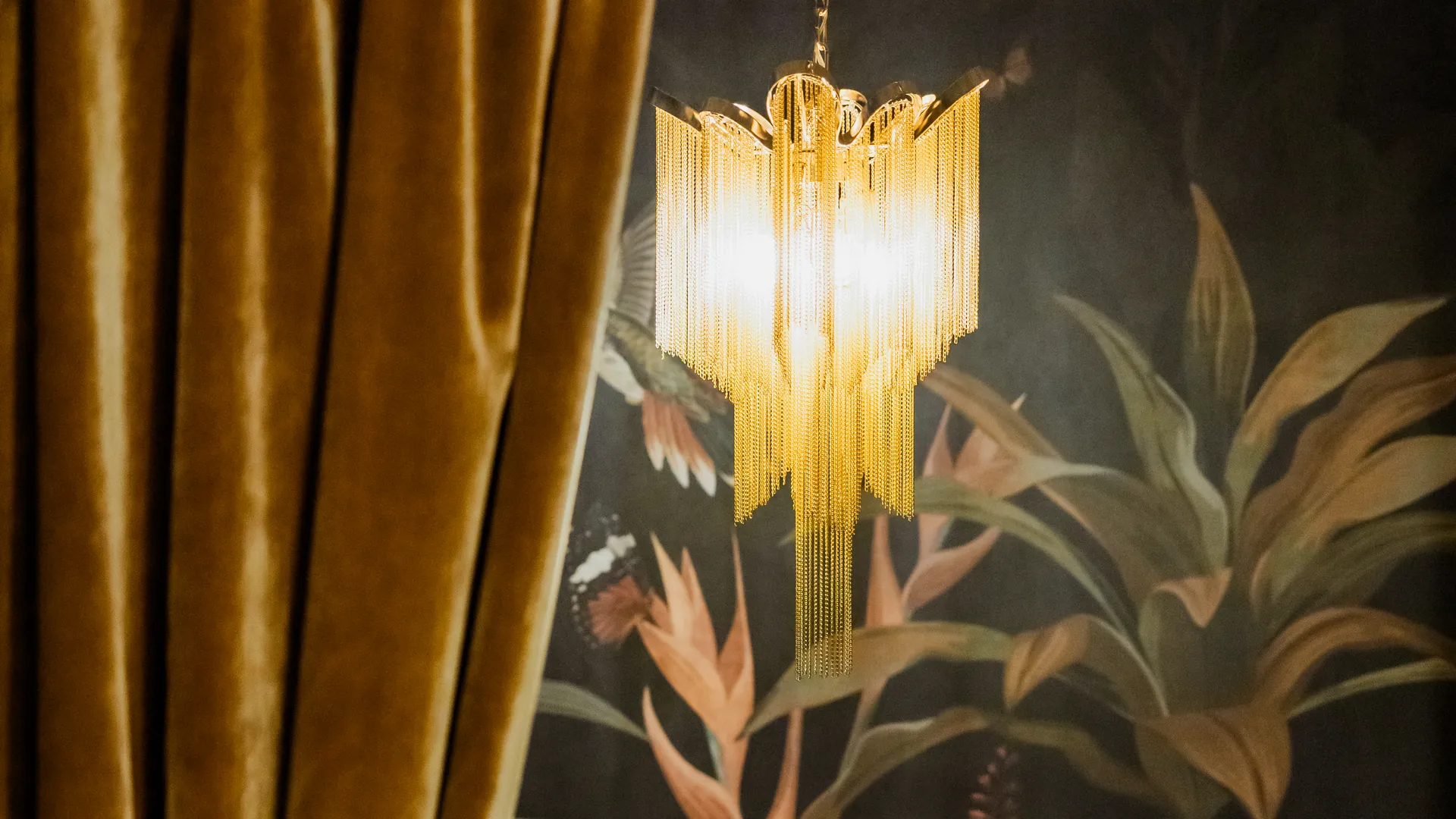 A drawn-back golden velvet curtain beside a glowing fringe chandelier inside Anigma