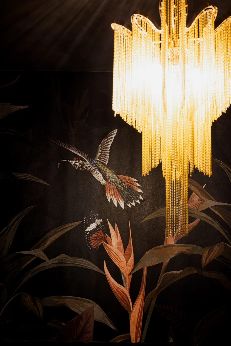 An amber chandelier glowing against botanical wallpaper at Anigma